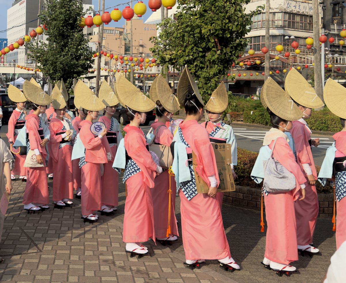 Tänzerinnen in rosa Yukata und großen Strohhüten stehen in einer Reihe auf einer Straße in Tokushima, über ihnen hängen rote und gelbe Laternen.