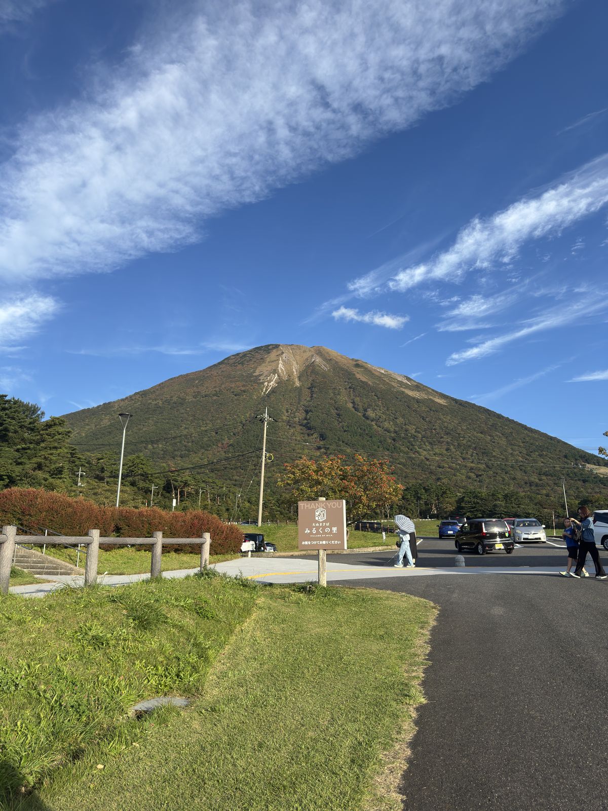 Der Daisen mit seinem spitzen Gipfel unter blauem Himmel, davor ein Parkplatz und ein Schild der Milchfarm Milk no Sato.